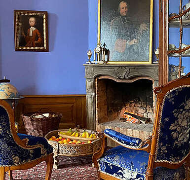 blaues Zimmer im Schloss, zwei Stühle mit blauem Samt bezogen vor einem Kamin, 2 Gemälde an der Wand, niedriger Beistelltisch dazwischen mit Äpfeln und Bananen 